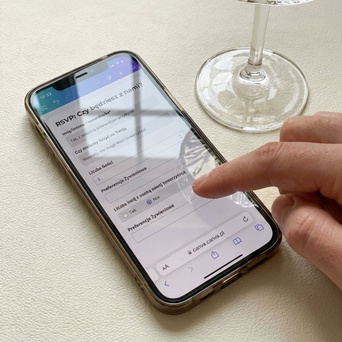 Dłoń obsługująca ekran smartfona z otwartym mobilnym formularzem RSVP na stronę weselną w przeglądarce, leżącym na jasnej fakturze obok kieliszka.