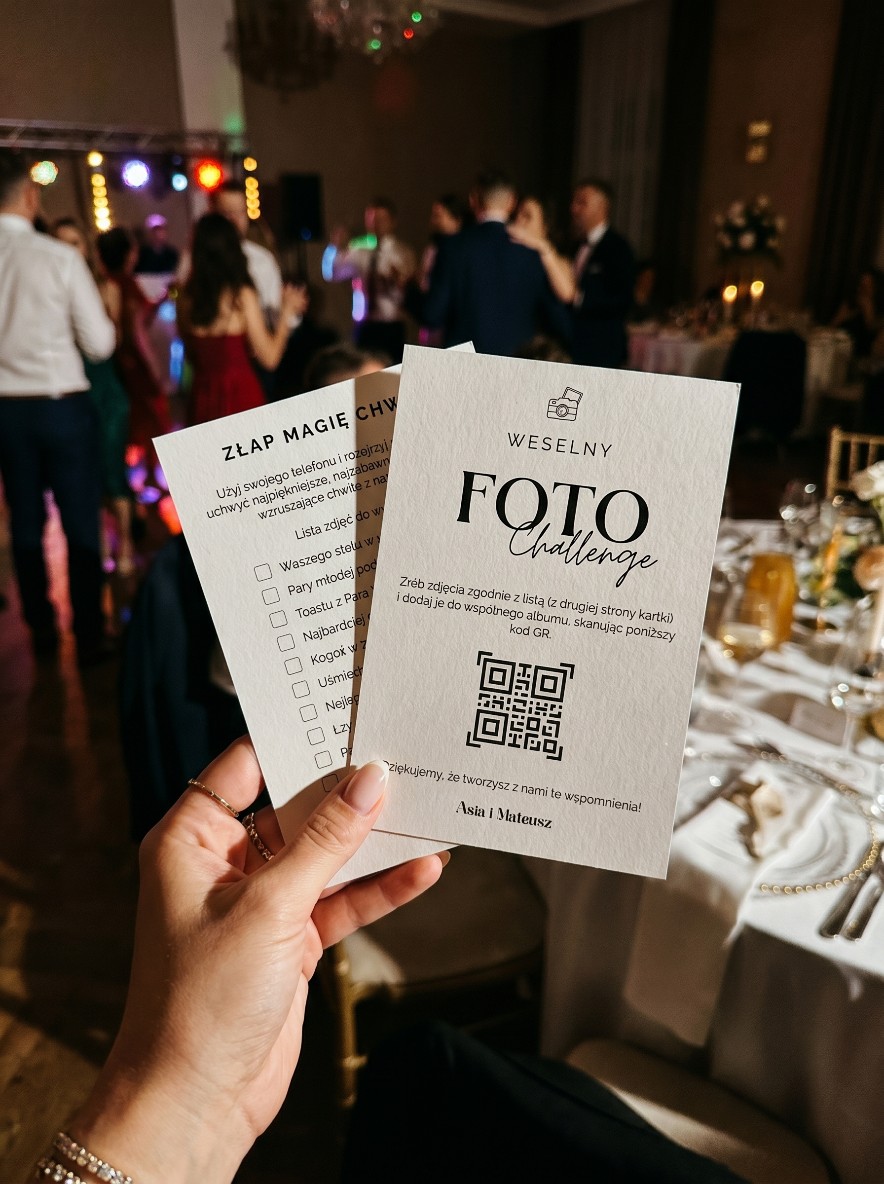 Dłoń trzymająca papierowe karty gry weselnej Foto Challenge na tle rozmytego, roztańczonego parkietu.